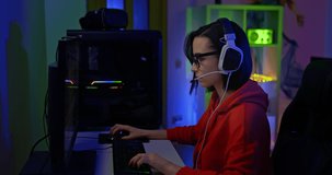 Girl sits at a computer in headphones with a microphone and plays a computer game over the network, e-sports. Beautiful girl gamer sits in a dark room and plays an online game. 4k, ProRes
