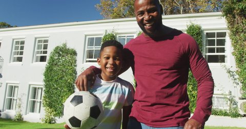 Portrait of african american dad and son smiling while holding a football in the garden. fatherhood and love concept