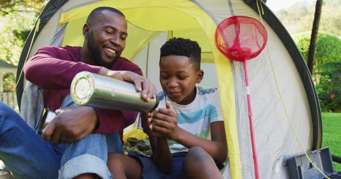 African american dad pouring coffee in his son's cup while sitting in a tent together in the garden. fatherhood and love concept