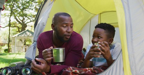 African american dad and son toasting and drinking coffee in a tent together in the garden. fatherhood and love concept