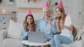 Smiling young adult mother, cute adorable child granddaughter and older mature grandmother in funny party hats congratulating relative birthday communicate video calling chatting by webcam app
