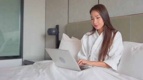 Asian woman in white bathrobe using her laptop computer sitting on the bed with her legs crossed at Hotel room, she is typing a message on laptop keyboard