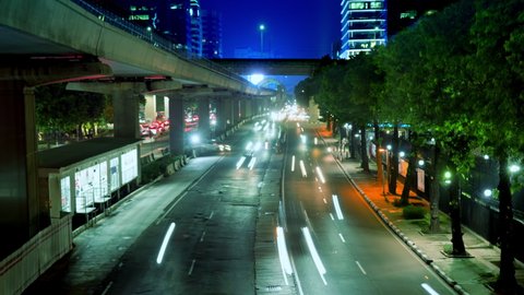 A beautiful timelapse of a night city road with neon glow of distant lightboxes and car lights. Urban scenery of dark street, trees, highway, automobiles, anonymous strangers passing by, colorful glow