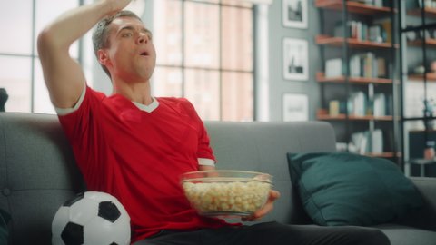 Handsome Young Adult Man Sitting on a Couch Watches Soccer Game on TV, Cheers for his Team to Win Championship. Joyful Fan Eats Snacks, Celebrates when Favourite Club Play. Medium Shot