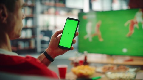At Home Soccer Fan Holds Green Screen Chroma Key Smartphone, Touching Screen, Watches Football Game on TV, Cheer for Favourtite Sports Team to Win Championship. Over the Shoulder