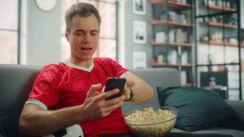 Charismatic Young Adult Man Sitting on a Couch Watches Game on TV, uses Smartphone App for Score, Bet, Statistics, Celebrates Victory when Team Wins Championship. Happy Fan Watches Sport. Medium Shot