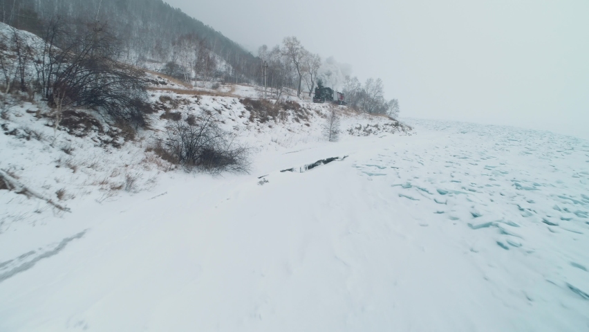 Unique aerial view Circum-Baikal railway incredible old steam locomotive train puffs of smoke from chimney goes fast through heavy snow storm snowfall blizzard near Baikal Siberia coastline. Russia 4k