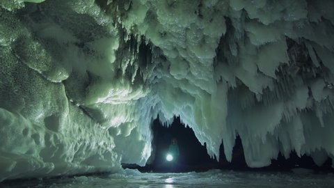 Impressive winter frozen cave splashes sharp ice floes of stalactites on ceiling. Man tourist at night comes out of dark grotto shines with flashlight illuminating walls pattern. Baikal Russia Siberia