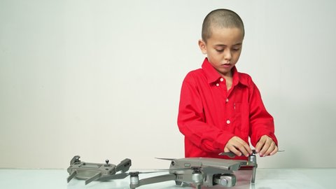 
Cute boy with skinhead hairstyle in a red long-sleeve shirt is assembling a drone 
on a clear glass table with a white backdrop.4K resolution.
