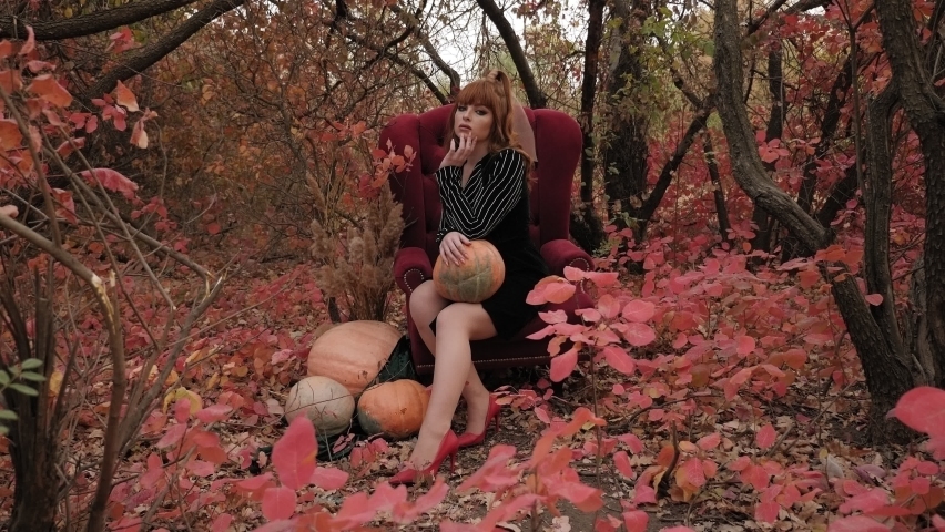 A red-haired model poses in the woods in autumn sitting in a red vintage chair.