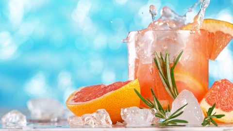 Super slow motion. Ice cube falling into glass of grapefruit gin cocktail or home made lemonade with sea surface background.