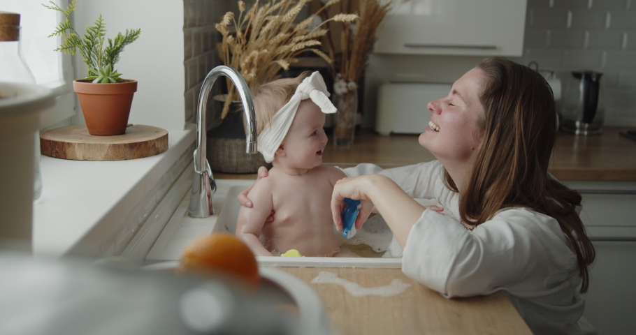 Mother and daughter spend time at home together. Young mom helps her adorable little child to take a bath in a kitchen sink. Adorable girl have fun in the water and foam. Royalty-Free Stock Footage #1074468797