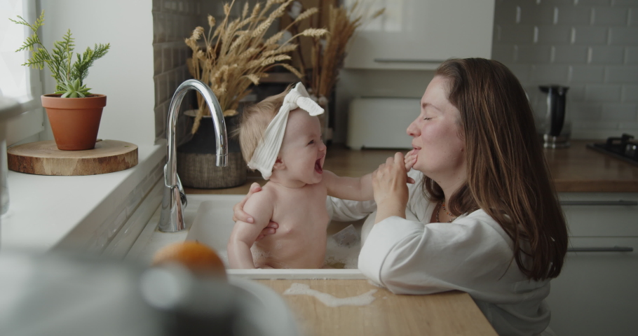Mother and daughter spend time at home together. Young mom helps her adorable little child to take a bath in a kitchen sink. Adorable girl have fun in the water and foam. | Shutterstock HD Video #1074468797