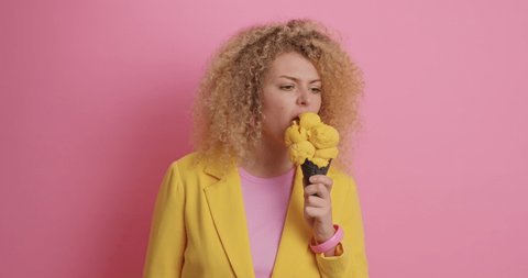 Curly haired young woman tastes new flavor of ice cream doesnt like it smirks face dressed in yellow jacket isolated over pink background. Dissatisfied female model eats tastless frozen dessert