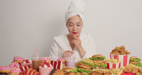 Happy young Asian woman chooses something delicious fast food eats fried chicken nuggets surrounded by harmful food isolated over white background feels hungry. Overdose and overeating concept