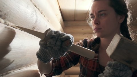 Young woman caulking and sealing wooden logs with oakum. Pretty woman works inside the wooden building and operates with hammer. Young feminist girl worker. Gender equality concept