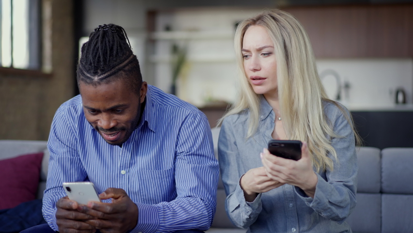 Jealous Caucasian wife looking at smartphone of African American husband talking and gesturing. Envious young beautiful woman arguing with unfaithful handsome husband messaging online in social media
