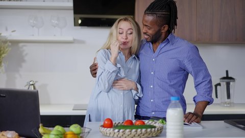 Pregnant Caucasian woman tasting organic ingredients for healthful salad as African American man hugging spouse talking in slow motion. Portrait of interracial couple cooking breakfast at home