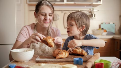 Smiling and laughing mother with little son eating freshly baked croissants on messy kitchen at home. Children cooking with parents, little chef, family having time together, domestic kitchen.