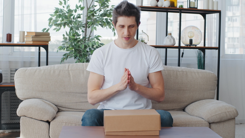 Caucasian man millennial customer buyer sitting on couch rubs palms waiting for gift opens unpacks cardboard box parcel hoping for good product feels disappointed sadness dissatisfaction with delivery Royalty-Free Stock Footage #1074477266