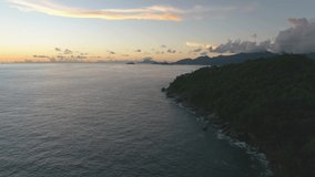 A drone flies over a rainforest in the Seychelles at pink sunset. The drone camera glides smoothly over the waves of the ocean. The ocean is visible on the horizon.