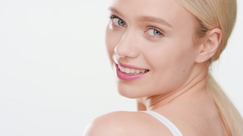 Attractive fit European young woman with long blond hair in ponytail in white crop top turns to the camera smiling wide for it on white background | Skin hydration concept