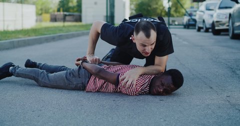Male police officer interrogating restrained African American man on asphalt ground during detention operation on street