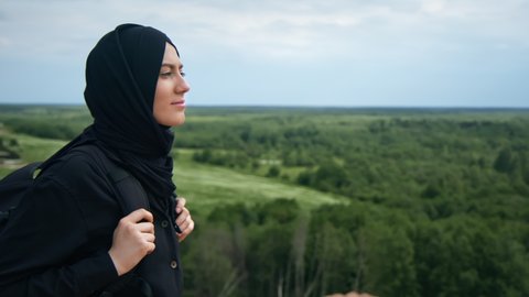 Smiling Muslim backpacker female admiring nature landscape from top of mountain enjoying summer travel vacation. Happy Saudi tourist woman contemplating scenery sky forest horizon freedom balance