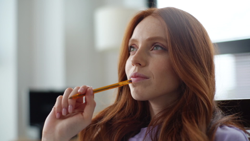 Close-up face of happy dreaming young woman making notes in paper notebook sitting on chair on background of window. Tracking shot of pretty lady writing down hopes and dreams of future into diary.