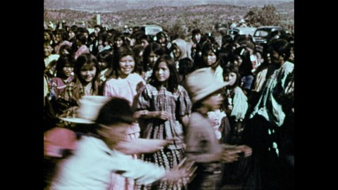 1970s: Groups of children chase after a girl. Girl resumes dancing. Dancers in headdress and hoods come in front of a group. Dancers move around in a circle.