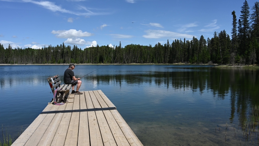 Gray haired man in shorts sitting on a bench on a wooden dock is fishing in a small lake.  
