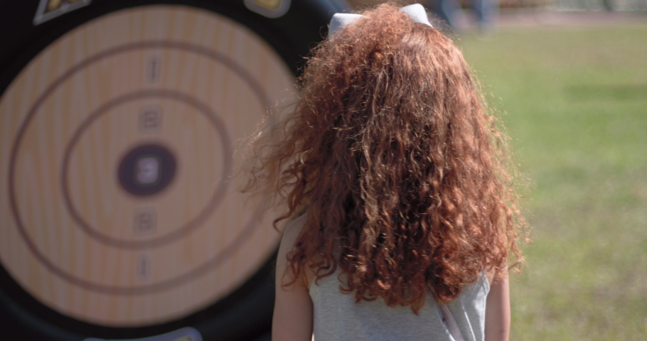 Cute young girl with red hair throwing toy axe at a target outdoors in a park midday. Filmed in 4k at 120fps.