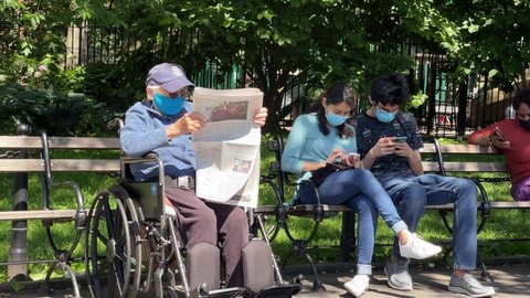 NYC, USA - JUNE 16, 2021: elderly person in wheelchair reading newspaper on bench in Washington Square Park next to people in mask New York City.