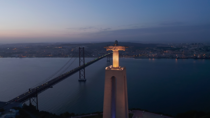 Aerial view of illuminated big Jesus statue above river spanned by cable-stayed bridge. Drone flying around Christ the King Sanctuary in Almada after sunset. Lisbon, capital of Portugal.