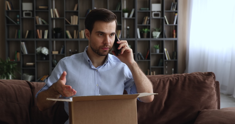 Man sitting on sofa with opened parcel express complaints to seller or courier on the phone about damaged goods, crashed items inside cardboard box. Dissatisfied e-commerce customer, claims concept Royalty-Free Stock Footage #1074542414