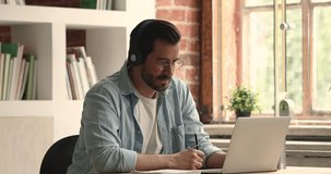 Happy young 30s caucasian man in eyeglasses wearing wireless headphones, watching educational online lecture, writing notes in copybook, enjoying distant learning, getting knowledge at home office.