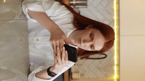 Portrait of excited redhead young woman getting surprisingly good online news on mobile phone sitting at table in light kitchen room. Surprised happy female celebrating victory on phone in apartment.