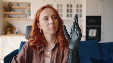 Young red-haired woman looks at her bionic prosthetic left hand and alternately flexes and unbends her fingers. Girl is adjusting the bionic prosthesis Bionics Cybernetic Robotic-arm Hand prosthesis.
