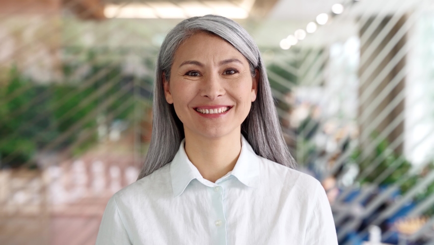 Happy confident smiling middle aged adult Asian older senior female businesswoman corporation ceo in modern office looking at camera. Business woman executive concept. Closeup portrait headshot. Royalty-Free Stock Footage #1074556382