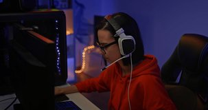 Portrait, a beautiful girl in glasses and headphones sits at a computer in a dark room. Streamer girl plays the game over the network and speaks over the microphone with the players. 4k, ProRes
