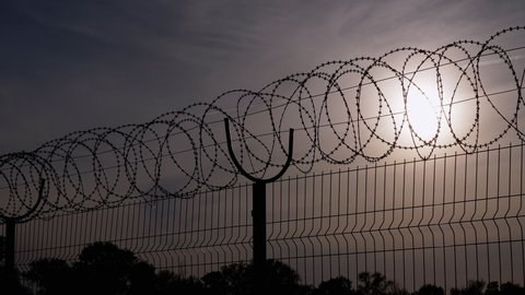Barbed Wire Hangs on Border of an Iron Fence Against the Backdrop of Sunset. Restricted area. Barrier from strangers people, refugees, prisoners. Lattice. Jail. 4K. Close up. Slow motion.