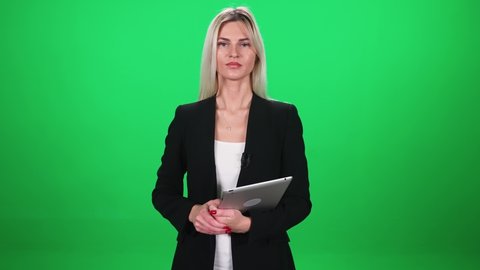 Woman reporter in suit looks into the camera and speaks in lavalier microphone, holding a tablet in his hands, female on a green background, template for TV news agencies, journalist at work
