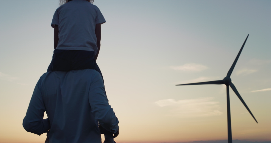 Back Shot of a Man Carrying his Child on His Shoulders and Walkingin a Wind farm. Happy Father and Daughter Enjoying a Beautiful Sunset and Clean Air in a More Environmental Friendly Life Royalty-Free Stock Footage #1074587402