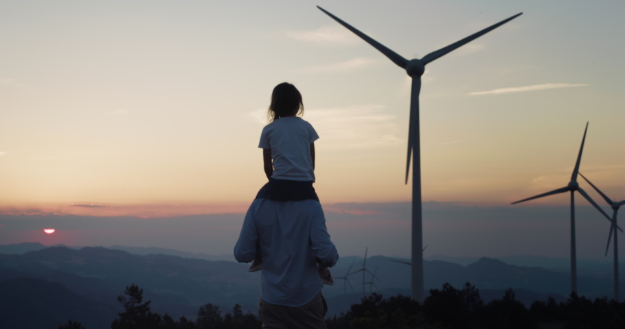 Back Shot of a Man Carrying his Child on His Shoulders and Walkingin a Wind farm. Happy Father and Daughter Enjoying a Beautiful Sunset and Clean Air in a More Environmental Friendly Life