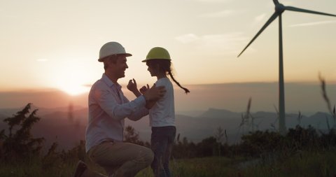 Cinematic shot of young father engineer putting on protective helmet to his little daughter on windmill field at sunset.Concept of renewable energy, love for nature, family, electricity, green, future