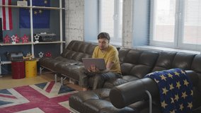 Handheld tracking shot of cheerful man with laptop sitting on couch in cozy apartment and beckoning his male partner and cute son to talk on video call with someone