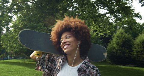 Cinematic shot of young african woman having fun to listening to music with earphones after skateboarding in green city park. Concept of lifestyle, youth, diversity, multiethnic, recreation, sports.