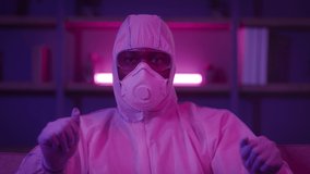 Unrecognizable Funny Black Man Dancing Wearing Protective Hazmat Suit, Glasses And Ffp Respirator Mask, Having Fun Celebrating End Of Pandemic In Dark Room Illuminated With Neon Light