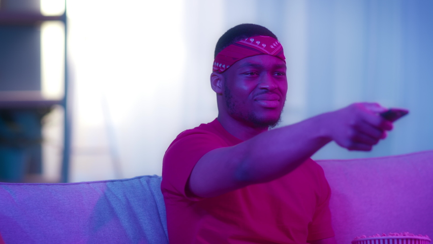 Discontented African American Guy Watching Sports On TV Pointing Remote Control Aside Switching Channels Sitting On Sofa At Home At Night. Displeased Viewer. Television Programming Problem
