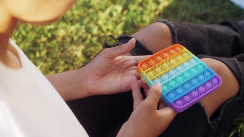 Latin teenager girl sitting on a grass at the street and playing pop it toy. Color silicone anti-stress toy pop it.